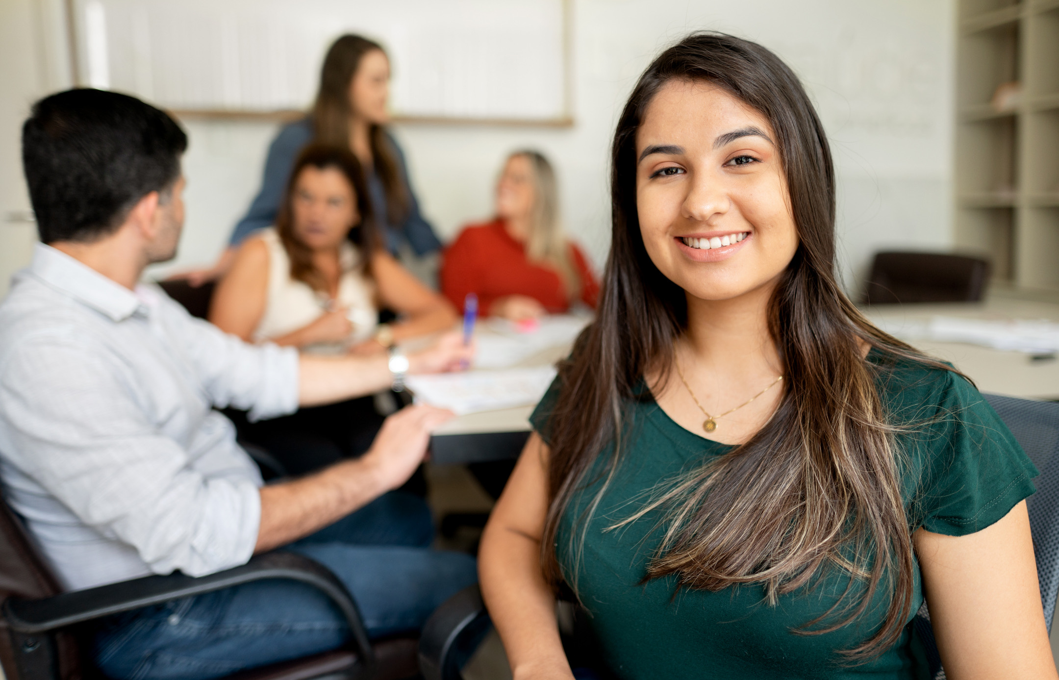 A young woman smiling at her desk, with colleagues meeting around a table behind her
